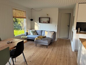 A cozy living room with a grey sofa, a wooden dining table, and a view of greenery outside.