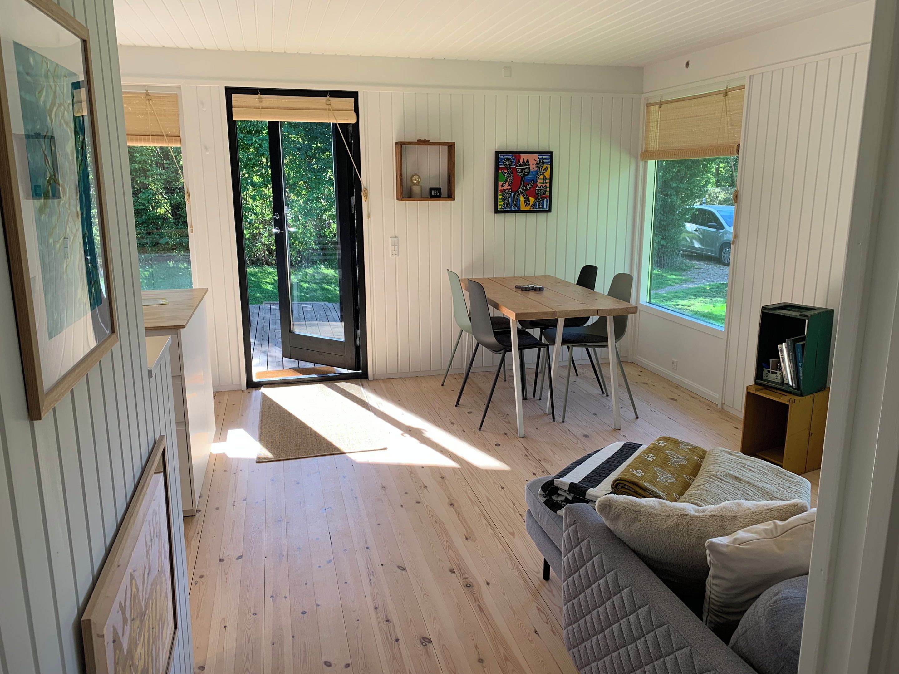 A room with a wooden floor, a dining table with chairs, a sofa, a bookshelf, and a door leading outside.