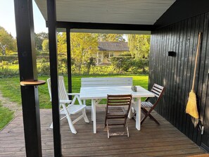 A covered outdoor seating area with a table and chairs, a broom leaning against the wall, and a view of a garden and house in the background.