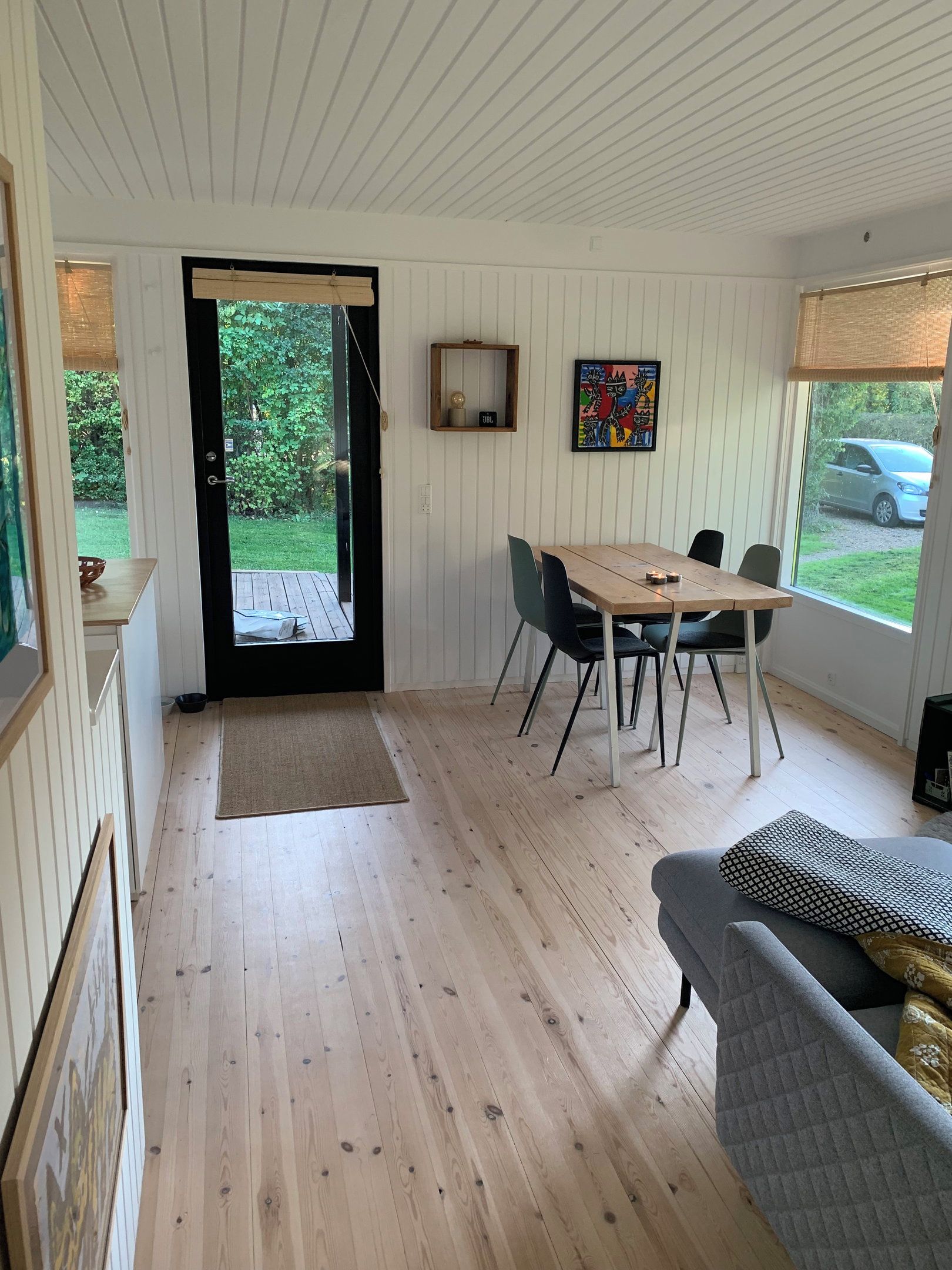 A room with a wooden floor, a dining table with chairs, a sofa, a door leading outside, and a window with a view of a car.