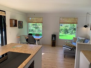 A room with a dining table, chairs, a sofa, a bookshelf, and large windows with a view of greenery.