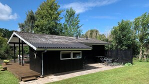 Een klein, donker houten tuinhuis met pannendak, een terras met tafel en stoelen, en een omheinde tuin.