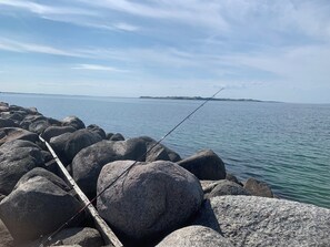 Una caña de pescar sobre rocas junto a un cuerpo de agua tranquilo, con tierra visible a lo lejos.