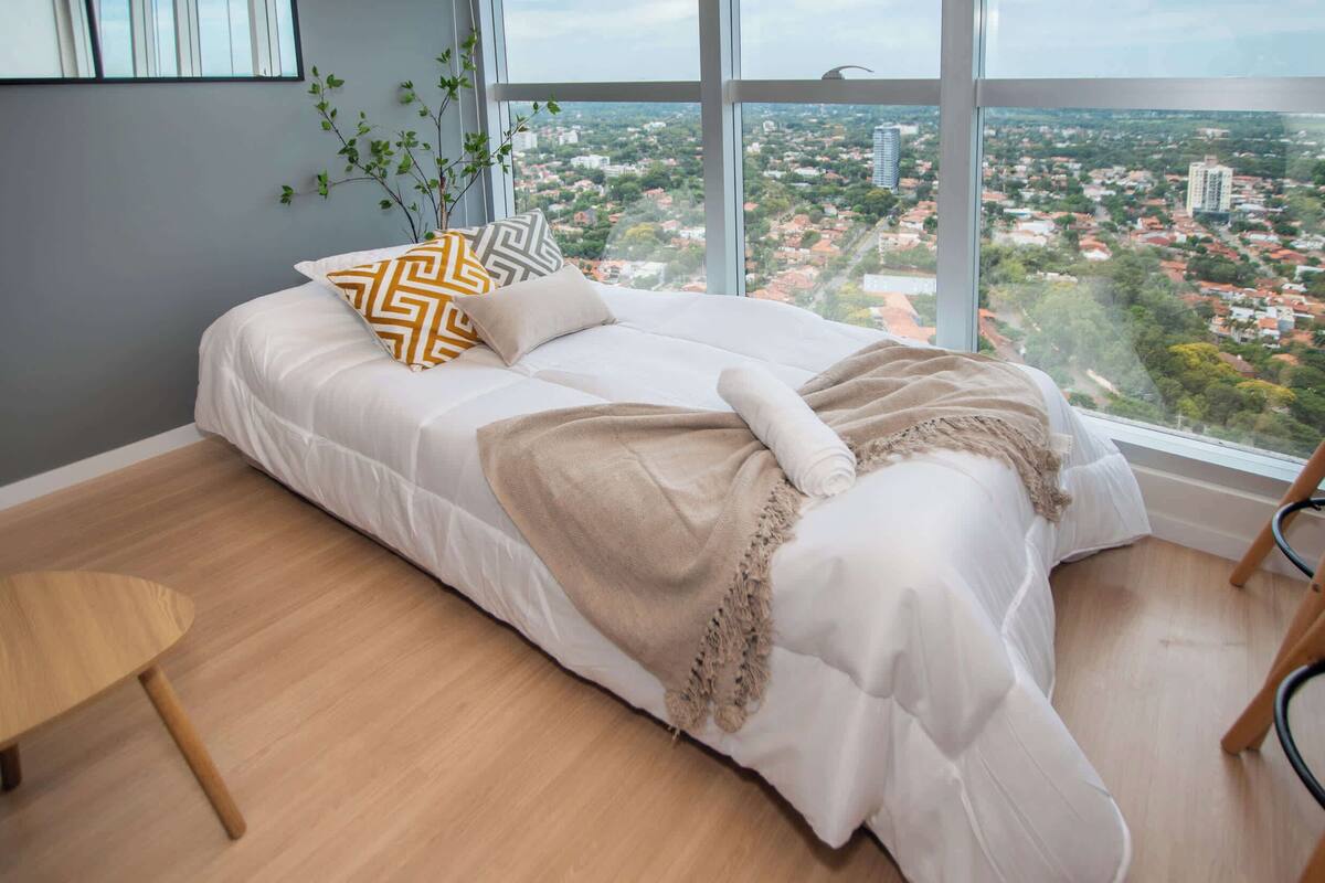 Ein Schlafzimmer mit einem Bett, Blick auf eine Stadtlandschaft, einem kleinen Holztisch und einer Pflanze.