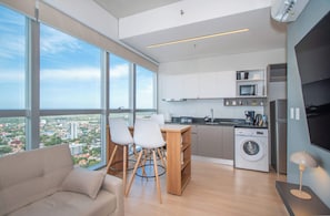 Eine moderne Wohnung mit großem Fenster, einer Couch, einem Esstisch mit Stühlen, einer Küche mit Geräten und einer Waschmaschine.