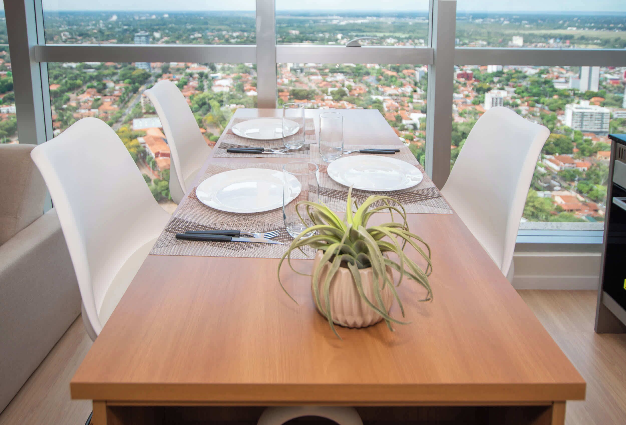 Uma mesa de jantar com quatro cadeiras, uma planta em vaso e vista para um horizonte urbano através de grandes janelas.