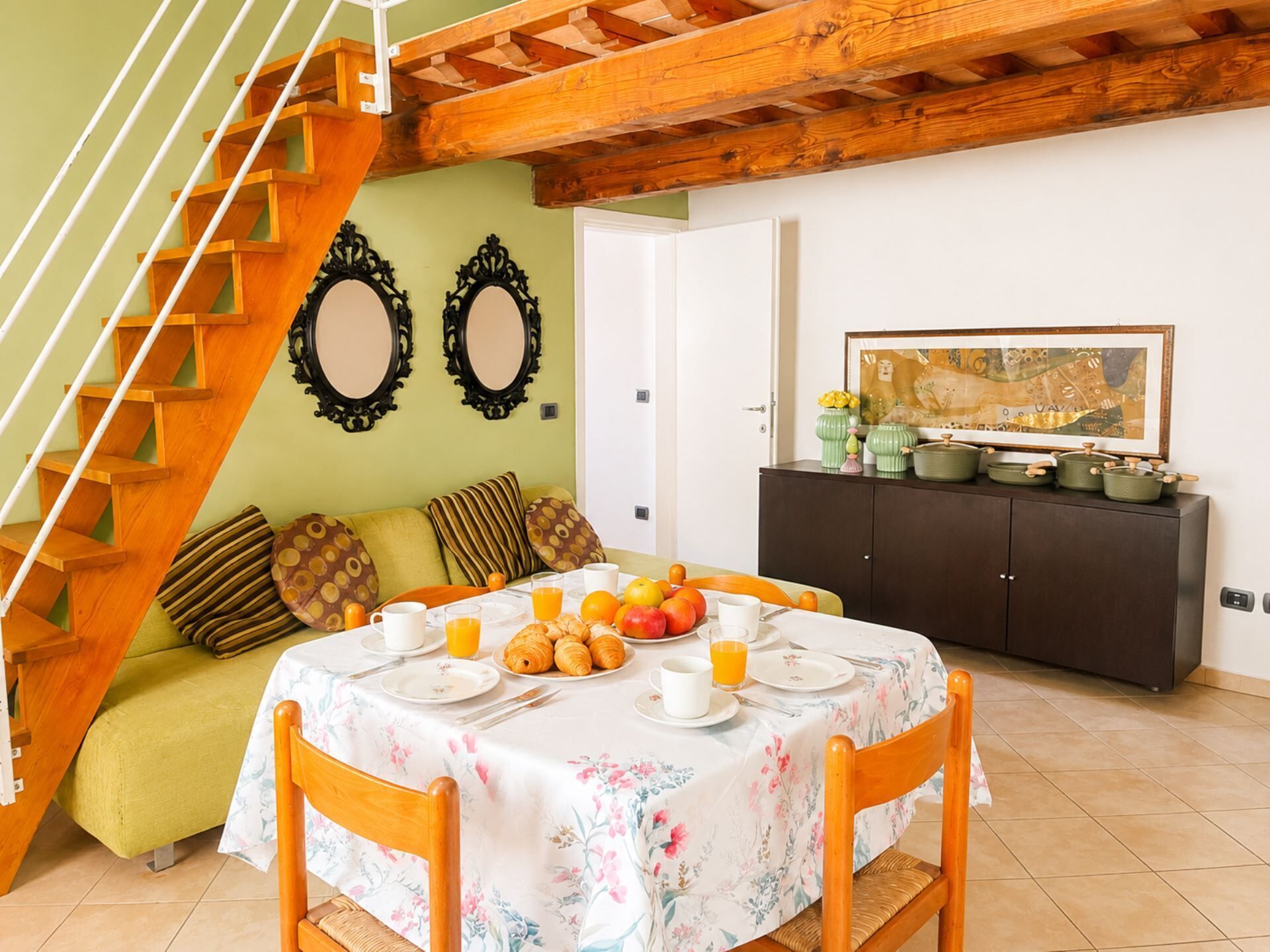 A dining area with a table set for breakfast, a staircase, a sofa, and a cabinet with kitchenware.