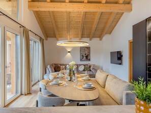 Un comedor moderno con techo de madera, una mesa redonda dispuesta para una comida y un sofá al fondo.