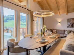 Ein Essbereich mit einem langen Tisch, der für eine Mahlzeit gedeckt ist, großen Fenstern mit Blick auf eine grüne Landschaft und einer Holzbalkendecke.