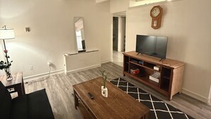 Una sala moderna con una mesa de centro de madera, un sofá negro, un televisor sobre un mueble de madera y una alfombra estampada.