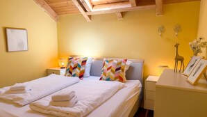 Una cama doble con sábanas blancas y almohadas de estampado color chevron, una mesita de noche con lámpara, y un pequeño escritorio con una figurita de jirafa y fotografías enmarcadas.