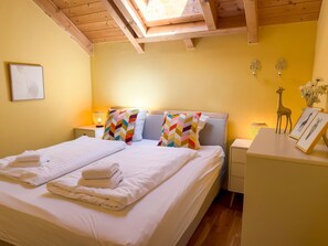 A double bed with white linens and colorful chevron-patterned pillows, a bedside table with a lamp, and a small desk with a giraffe figurine and framed pictures. - Chalet with nice garden and wood-fired hot tub (Kötschach-Mauthen)