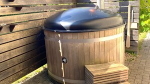 Una estructura de madera al aire libre con cubierta metálica, posiblemente una parrilla o un depósito, junto a un banco de madera y una pila de tablones.