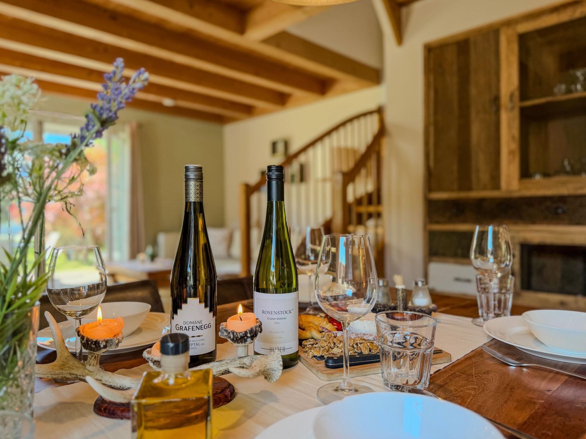 Une table à manger dressée avec des bouteilles de vin, des verres et une bougie, dans une pièce dotée de poutres en bois et d’un escalier.