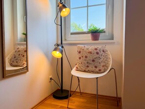 Quarto de canto com abajur, poltrona com almofada florida, janela com planta e espelho refletindo o ambiente.