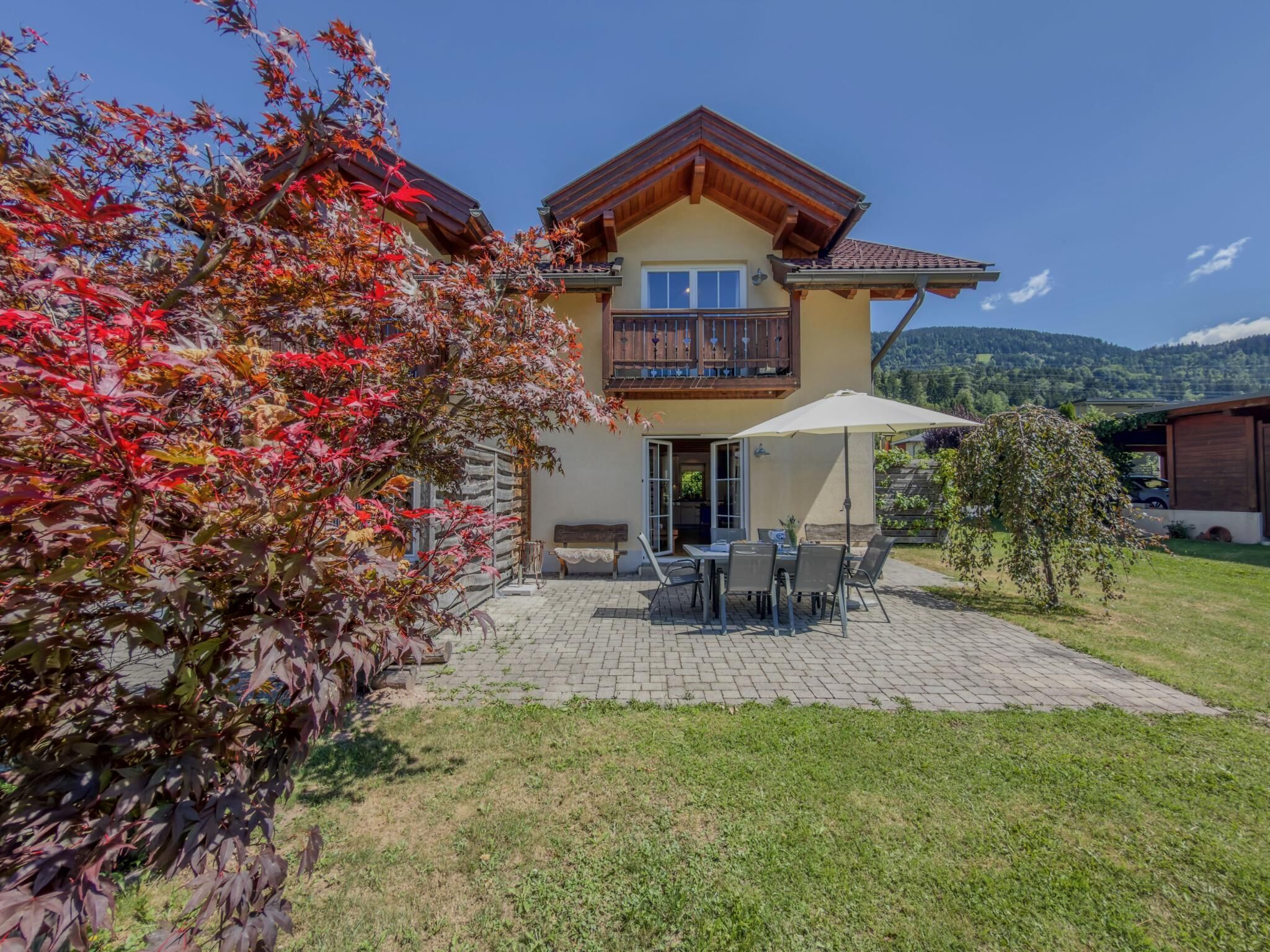 A two-story house with a balcony, a patio with a table and chairs, and a garden with trees.