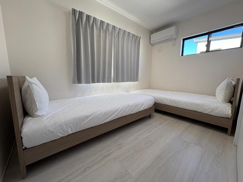 A room with two beds, a window with curtains, an air conditioning unit, and a light-colored wooden floor.