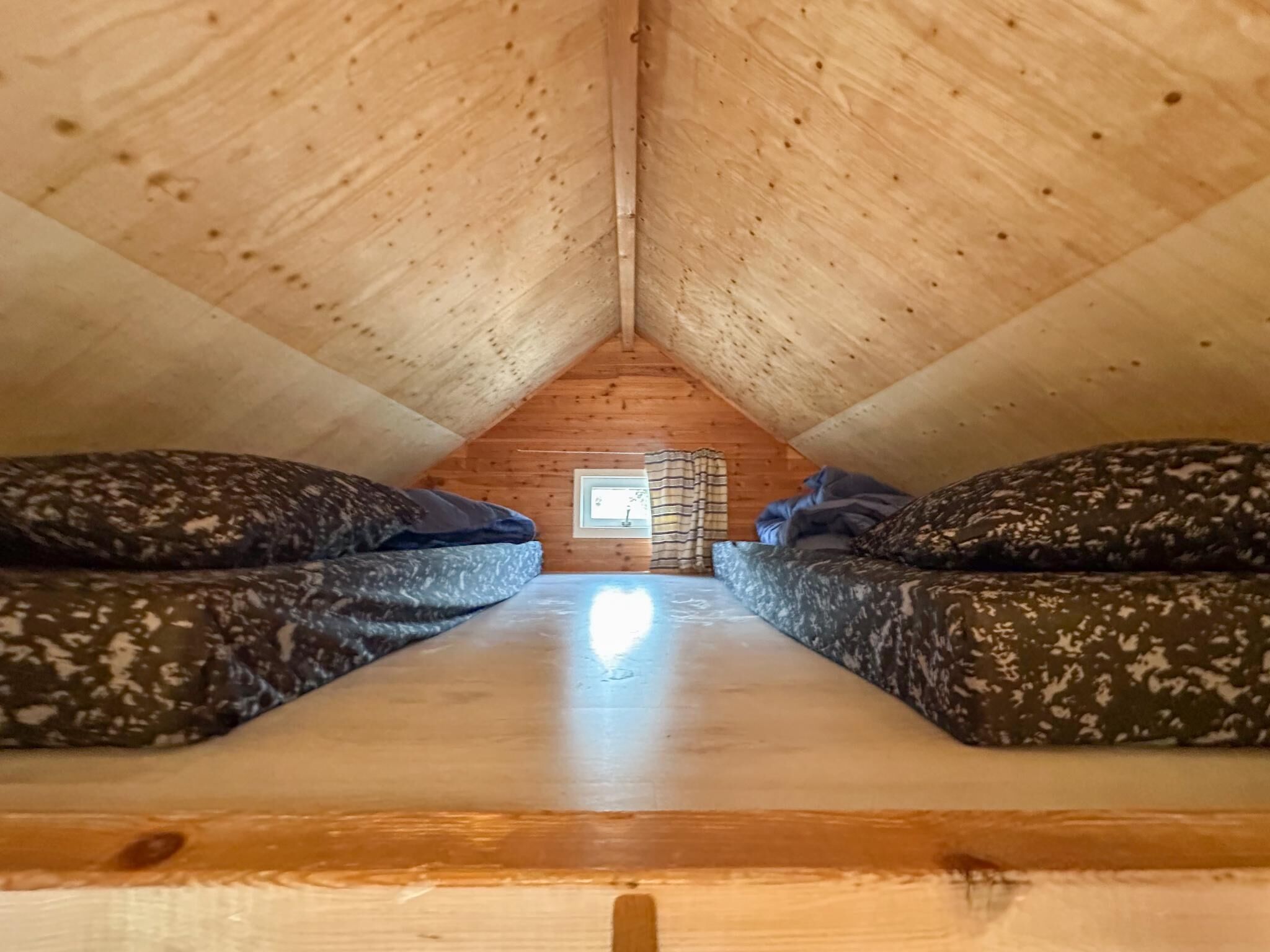 An attic bedroom with a sloped ceiling, two camouflage-patterned beds, a small window, and a wooden floor.