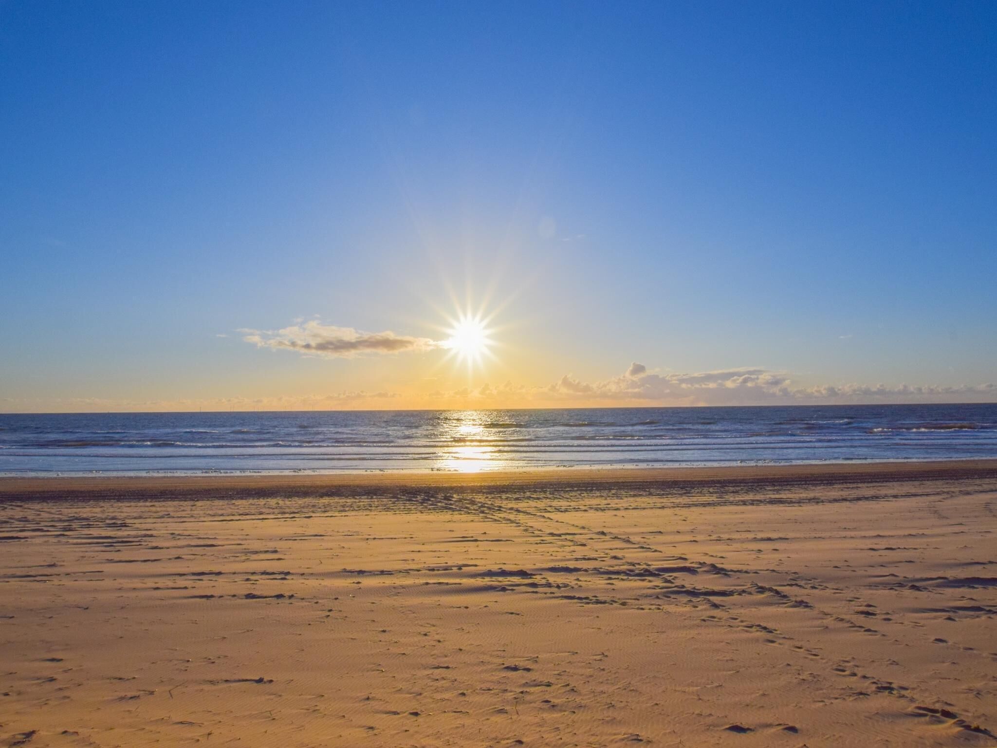 A beach with the sun setting over the horizon, calm sea, and sandy shore.