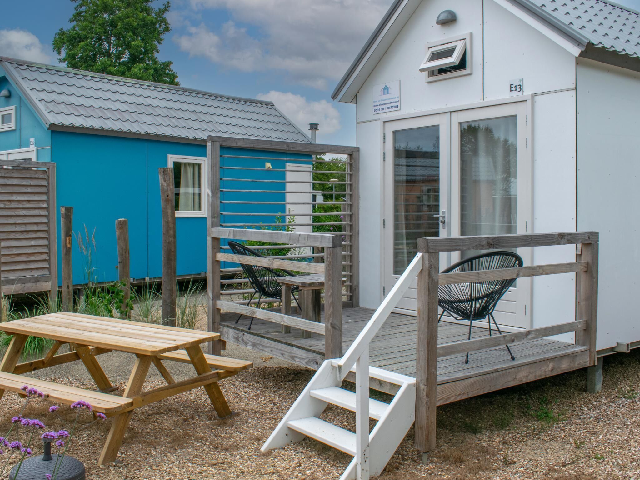 A small, single-story building with a white exterior and a blue exterior. There is a wooden deck with a bench and a table, and a set of stairs leading up to the deck.