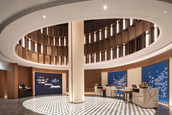 A modern hotel lobby with a central atrium, reception desk, and a digital welcome screen.