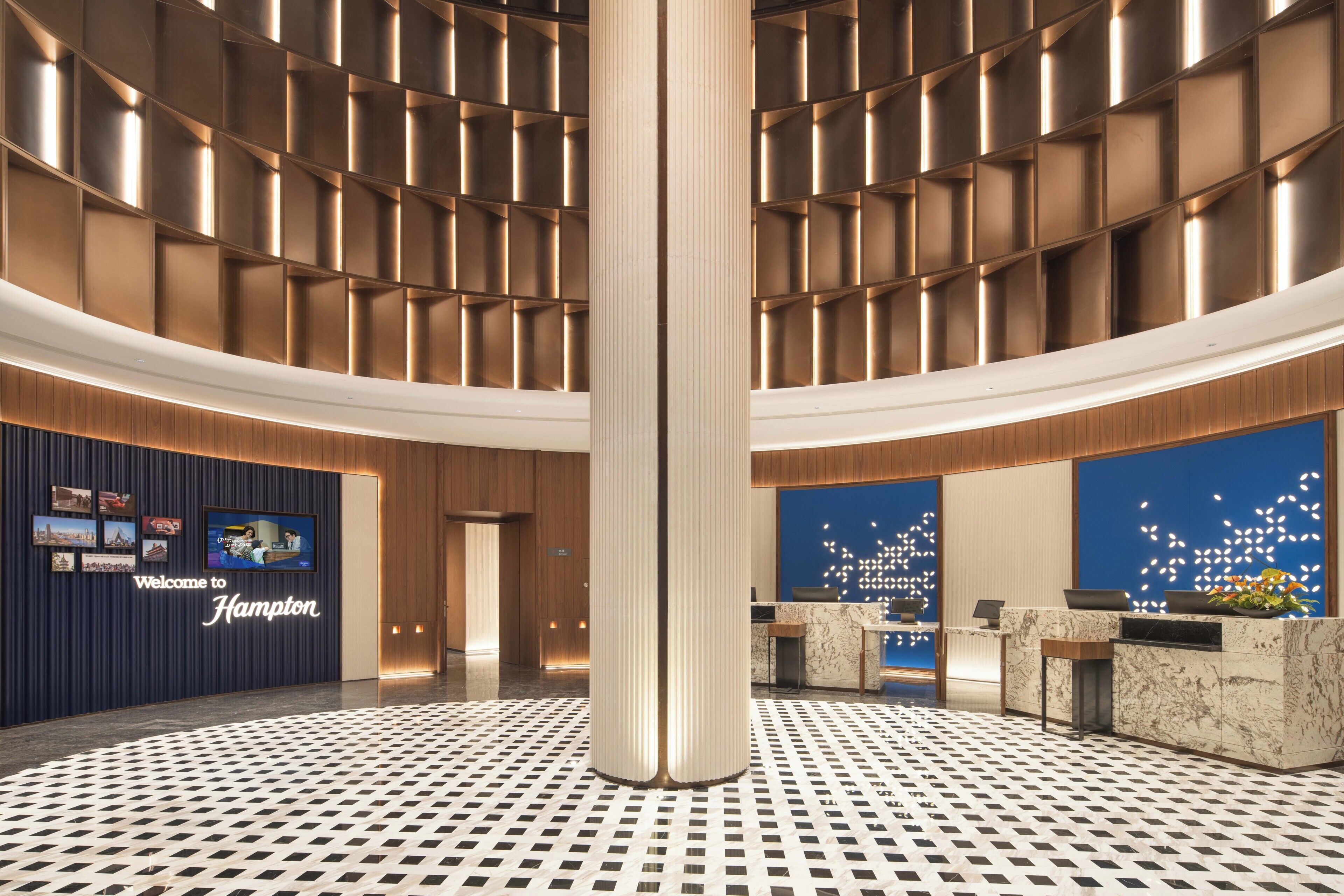 Un moderno vestíbulo de hotel con una columna central, mostrador de recepción y una pantalla digital mostrando un mensaje de bienvenida.