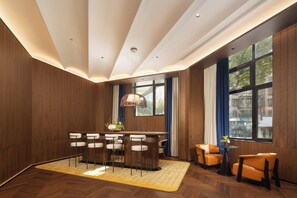 A modern hotel room with a wooden interior, a dining area with bar stools, a sofa, and a large window offering a view of the city.