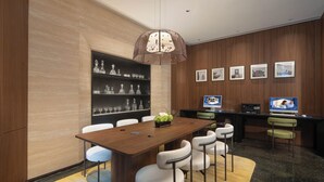 Un salón de conferencias moderno con una mesa de madera, sillas blancas, una lámpara colgante decorativa, una estantería con cristalería y cuadros en la pared.