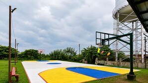 An outdoor basketball court with a hoop, surrounded by grass and trees.
