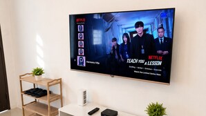 A modern living room with a flat-screen TV mounted on the wall, a white entertainment unit with shelves, and a small wooden shelf with a plant.