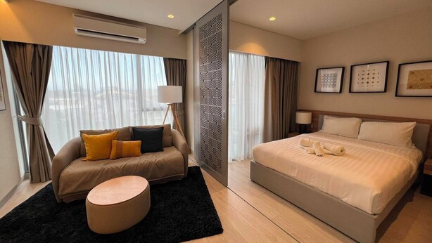 A modern hotel room with a bed, a sofa, a round ottoman, and a large window with curtains. - Laguna Lakeside Residences Phuket -YSLL (Tambon Si Sunthon)