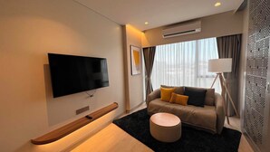 A modern hotel room with a flat-screen TV, a sofa with cushions, a round ottoman, a standing lamp, and a patterned wall panel. - Laguna Lakeside Residences Phuket -YSLL (Tambon Si Sunthon)
