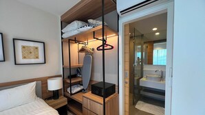 A hotel room with a bed, a bedside table with a lamp, a wardrobe with hanging clothes, a microwave, and a glass door leading to a bathroom with a sink and mirror. - Laguna Lakeside Residences Phuket -YSLL (Tambon Si Sunthon)