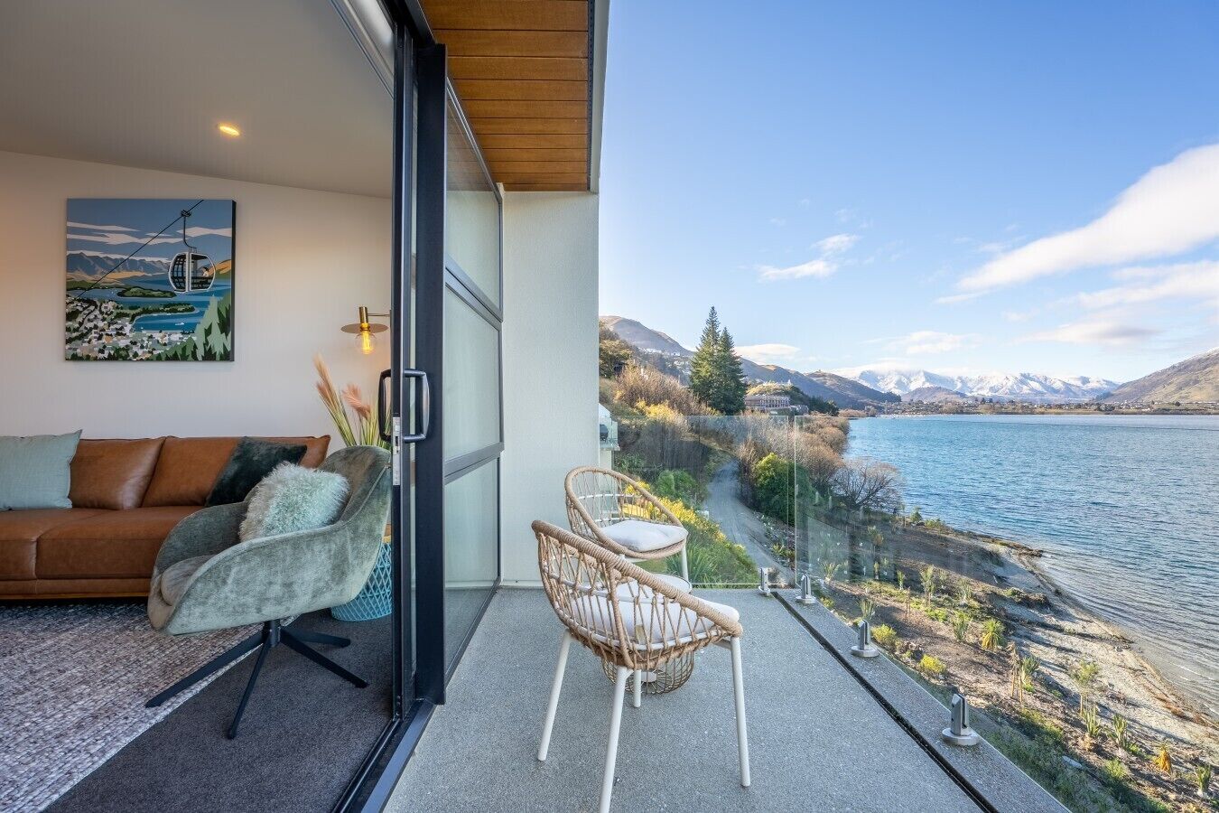 A balcony with a sofa and chairs, a glass door, and a view of a lake and mountains.