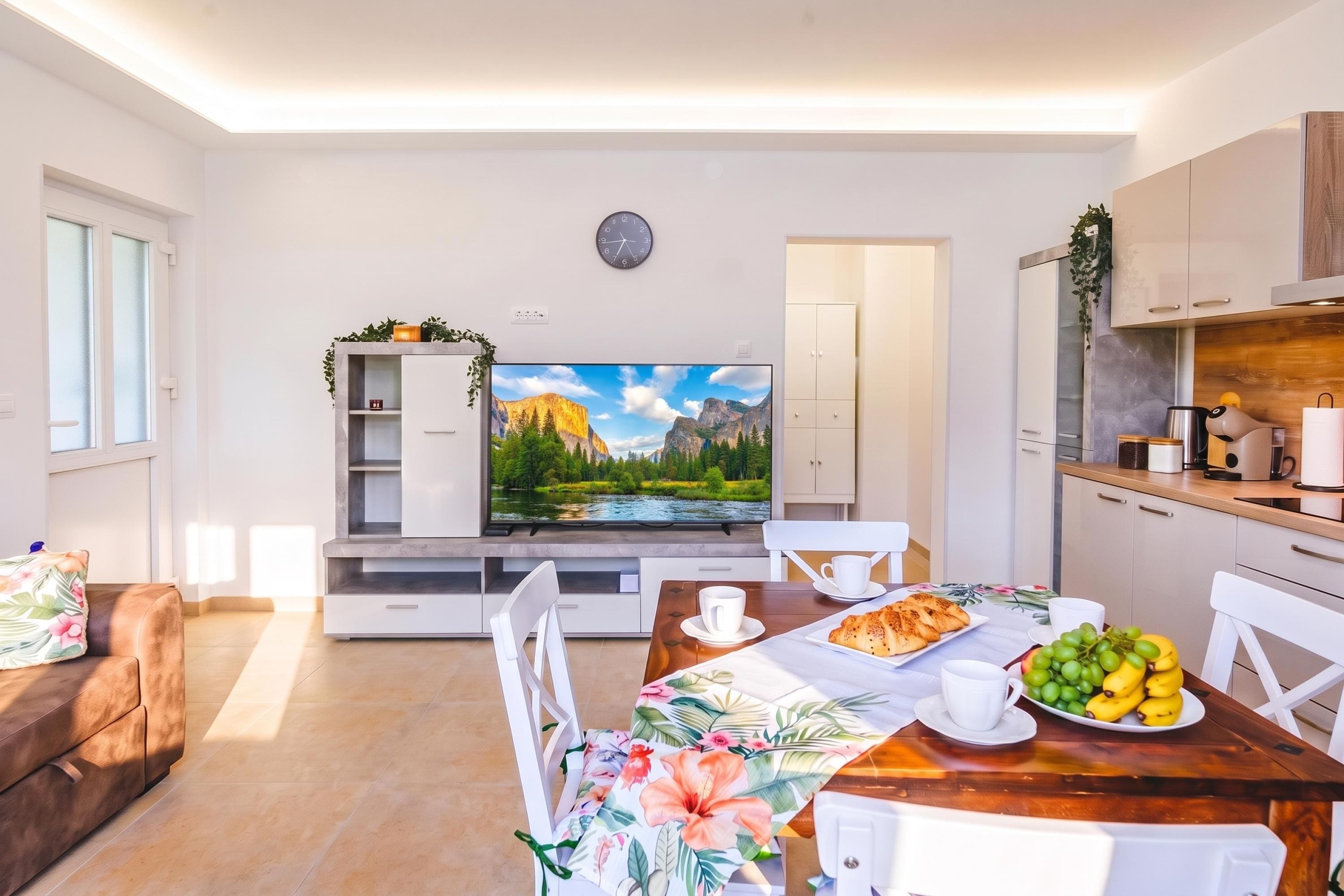 A modern living room with a flat-screen TV, a dining table with chairs, a sofa, and a kitchen area with cabinets and appliances.