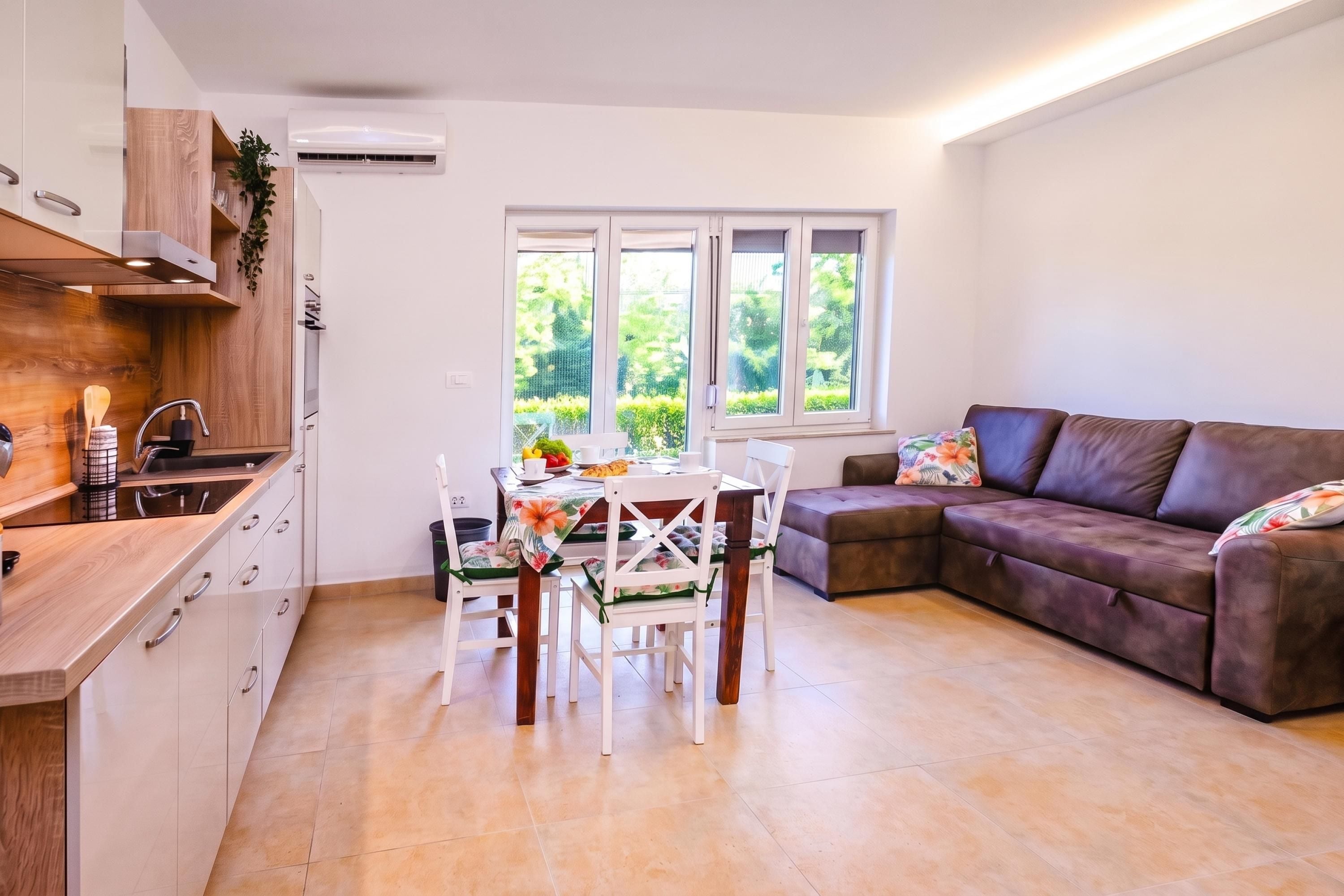 A modern kitchen with white cabinets and a wooden countertop, a dining area with a table and chairs, and a brown leather sofa with a floral cushion.