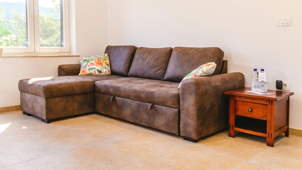 A brown sectional sofa with floral cushions, a wooden nightstand with a drawer, and a window with a view of greenery. - Sanbaldo 1BR Terrace Apartment Koper (Koper)