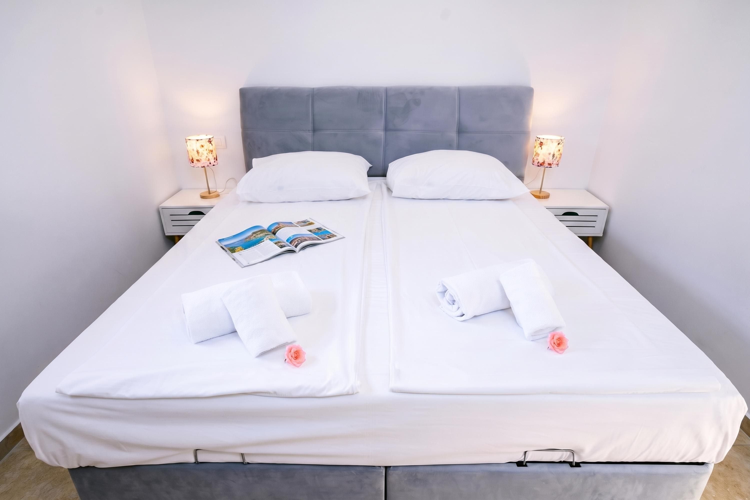 Ein Doppelbett mit weißen Laken, zwei Kissen, eine Zeitschrift und zwei aufgerollte Handtücher.