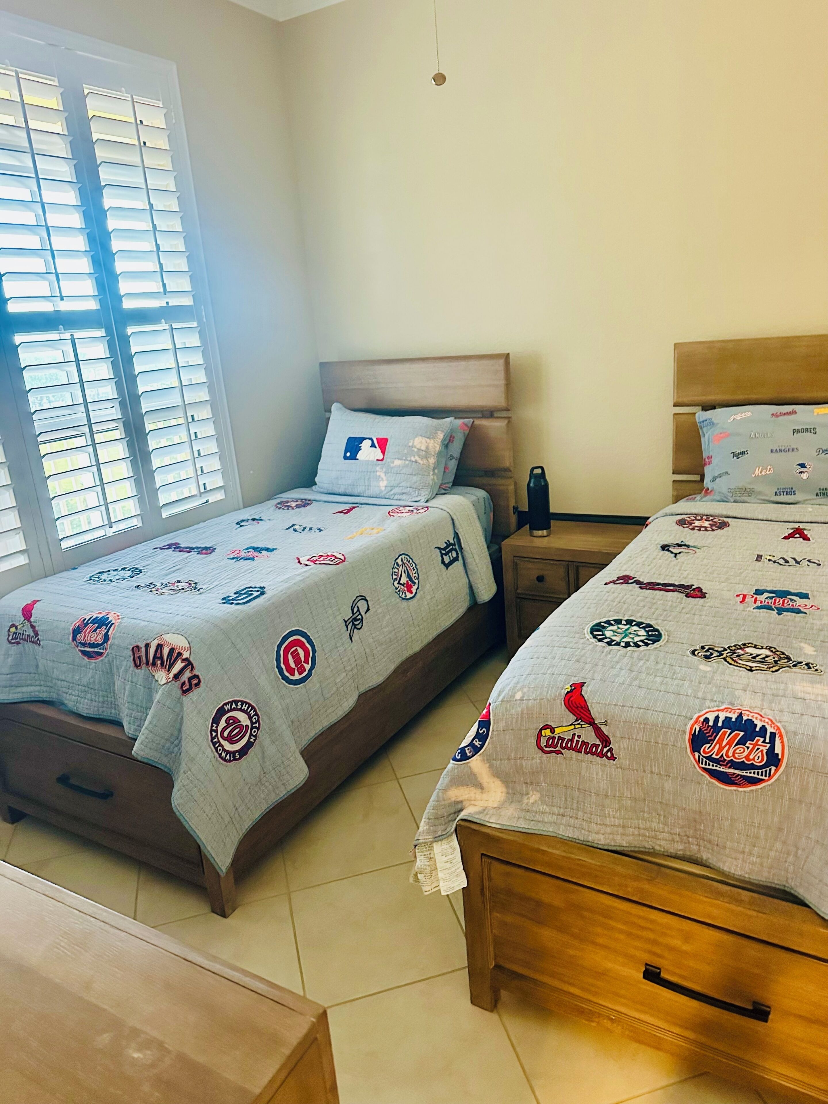Dos camas con ropa de cama temática de equipo de la MLB, un cabecero de madera, una mesita de noche con una botella de agua y una ventana con persianas.
