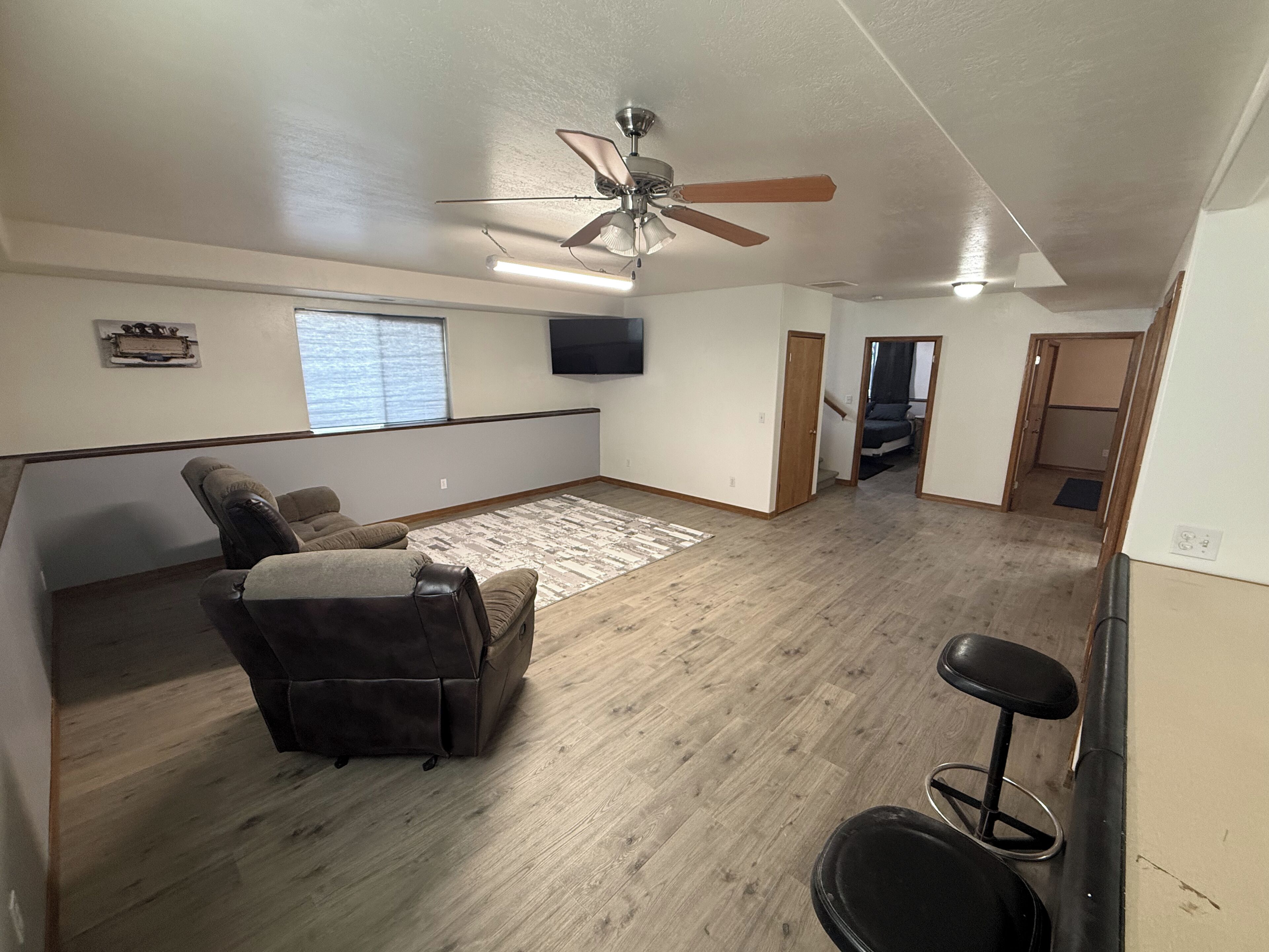A spacious living area with a ceiling fan, a flat-screen TV, a large window, and a wooden floor.