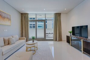 A modern living room with a sofa, coffee table, TV, and large windows. - Cozy 1 Bedroom l Burj Views from Pool Areal (Business Bay)