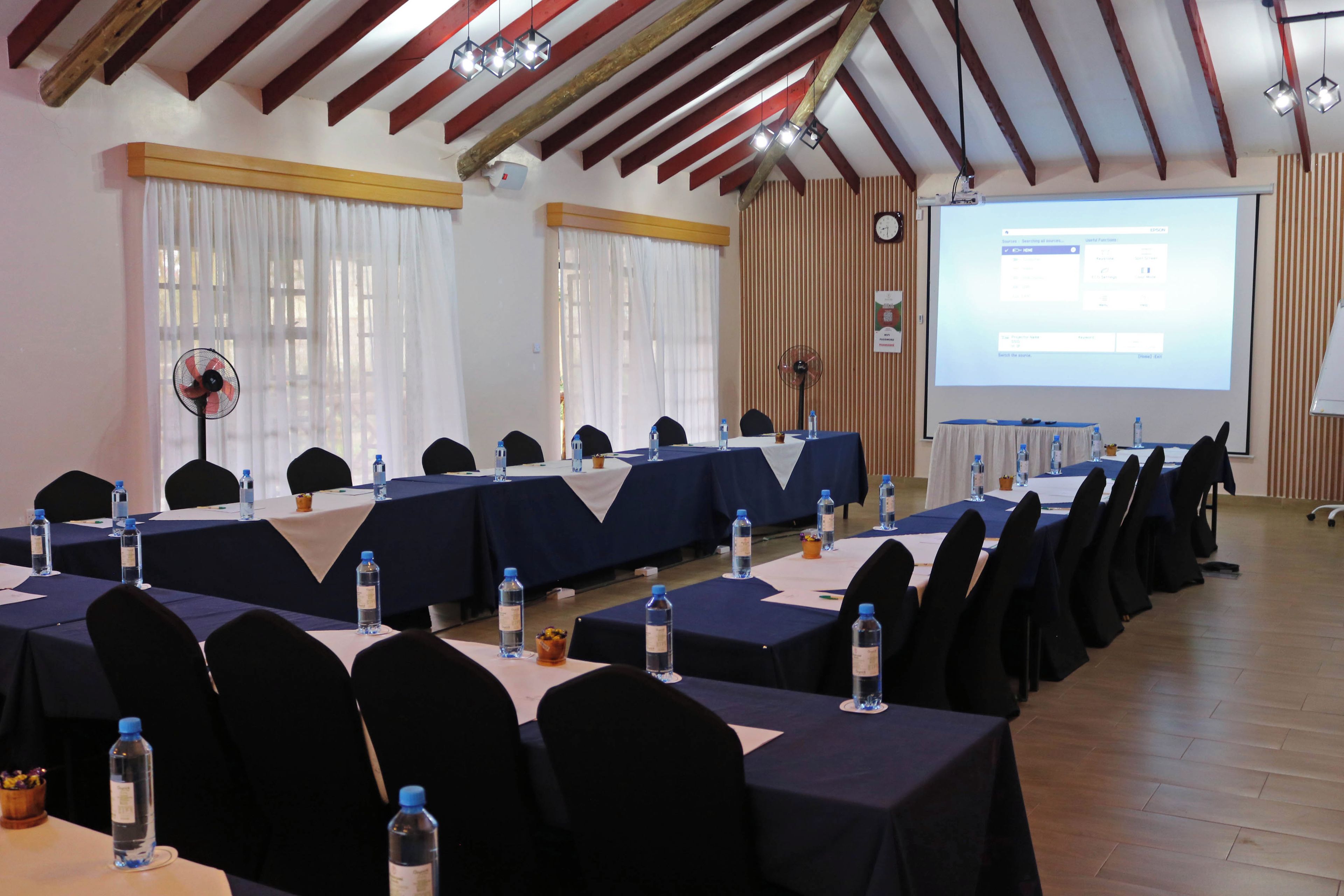Un salón de conferencias con largas mesas, sillas, botellas de agua y una pantalla de proyector mostrando una presentación.