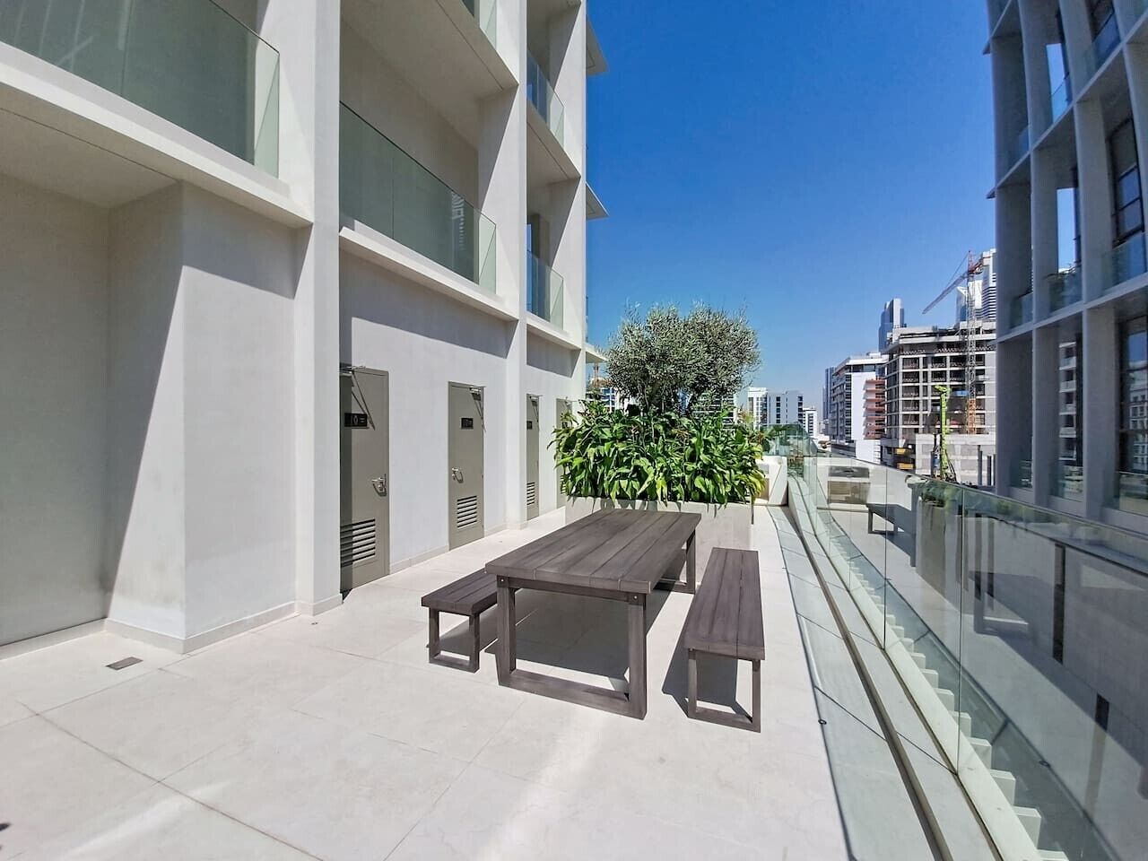 Eine moderne Außenpatio mit einem Holztisch und Bänken, umgeben von einem Glasgeländer und einem Gebäude mit Balkonen.
