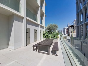 Un patio moderno al aire libre con una mesa y bancos de madera, rodeado por una barandilla de vidrio y un edificio con balcones.