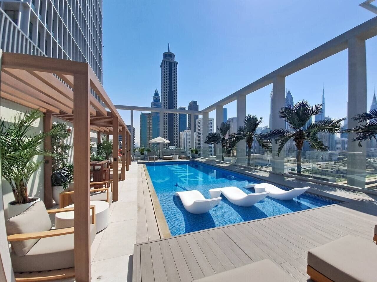 Une piscine sur le toit, avec des chaises longues, des palmiers et une vue sur les toits de la ville en arrière-plan.