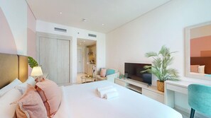 Quarto de hotel com cama, televisão, escrivaninha, cadeira, planta e banheiro visível ao fundo.