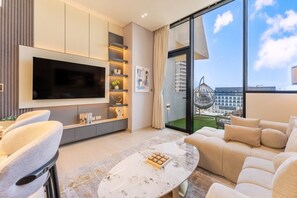 A modern living room with a large flat-screen TV, a sofa, a coffee table, and a view of a cityscape through large windows. - Luxe 1 Bed with Private Pool l Tennis Court (JVC)