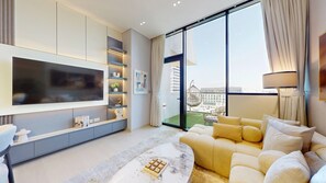 A modern living room with a large TV, a sofa, a coffee table, and a view of the city through large windows. - Luxe 1 Bed with Private Pool l Tennis Court (JVC)