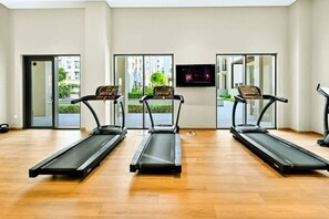 Ein Fitnessstudio mit drei Laufbändern, einem Bildschirm und großen Fenstern mit Blick auf Gebäude und Grünflächen.