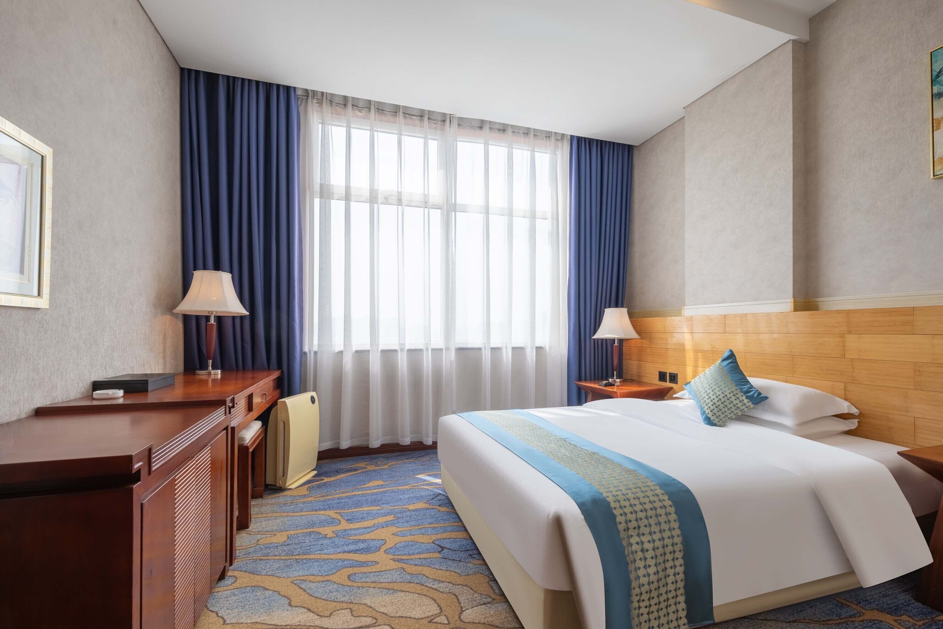 A hotel room with a bed, a desk, a chair, a lamp, a window with curtains, and a carpet.